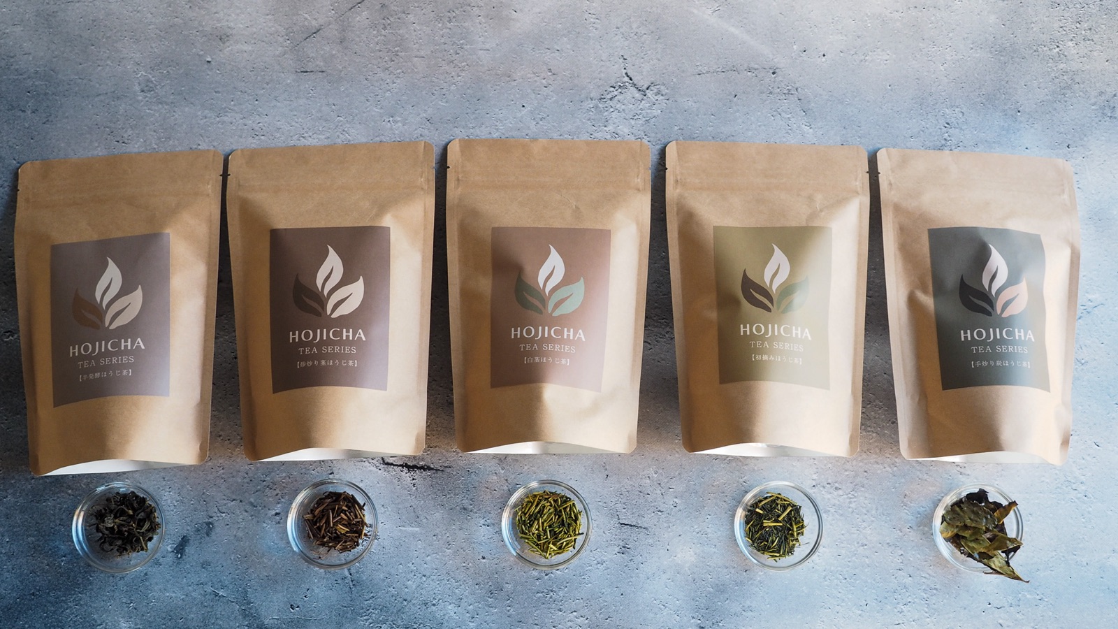 The tea bag packages of the five mirume hojicha in a row, each with a small glass dish of its dry tea.