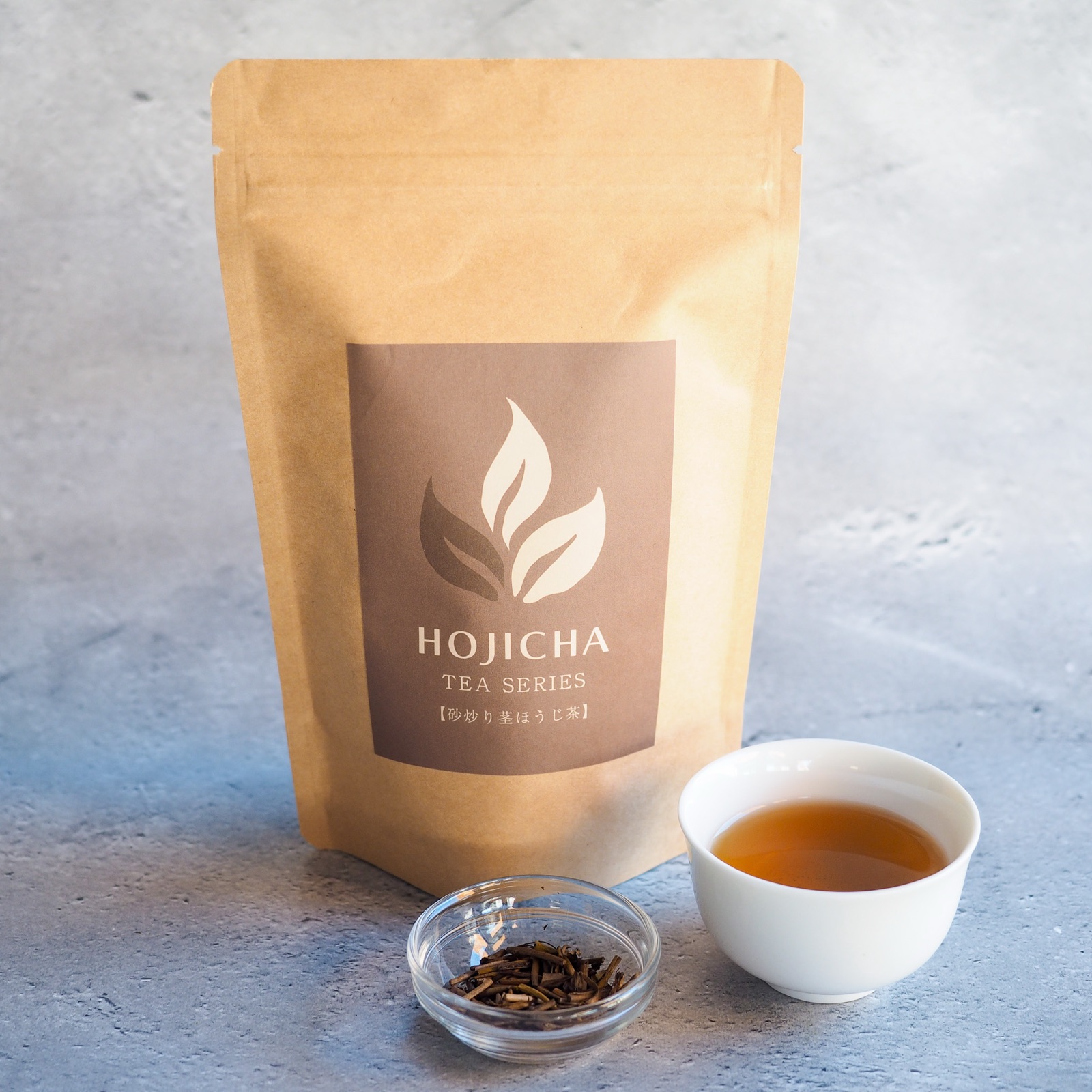 The tea bag package of No.1 Sand-Roasted Stem Hojicha, with a small glass dish of the dry tea and a white cup of the brewed tea.