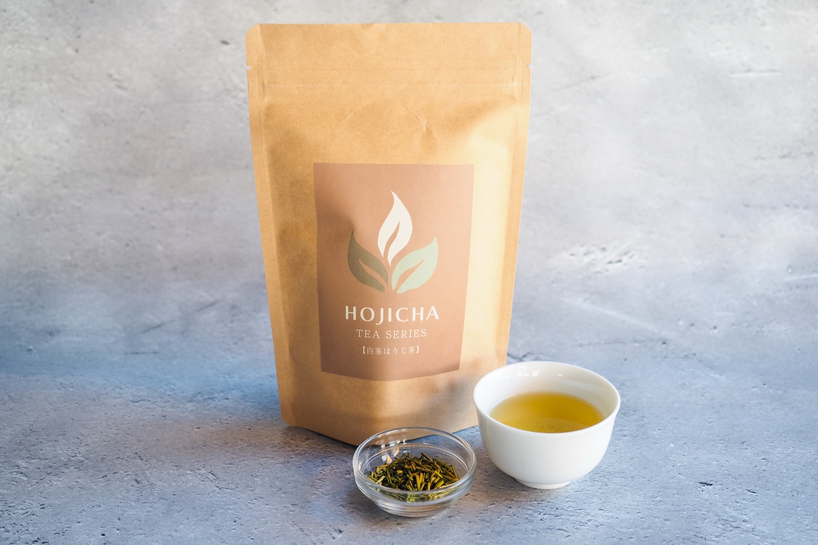 The tea bag package of No.2 White Stem Hojicha, with a small glass dish of the dry tea and a white cup of the brewed tea.