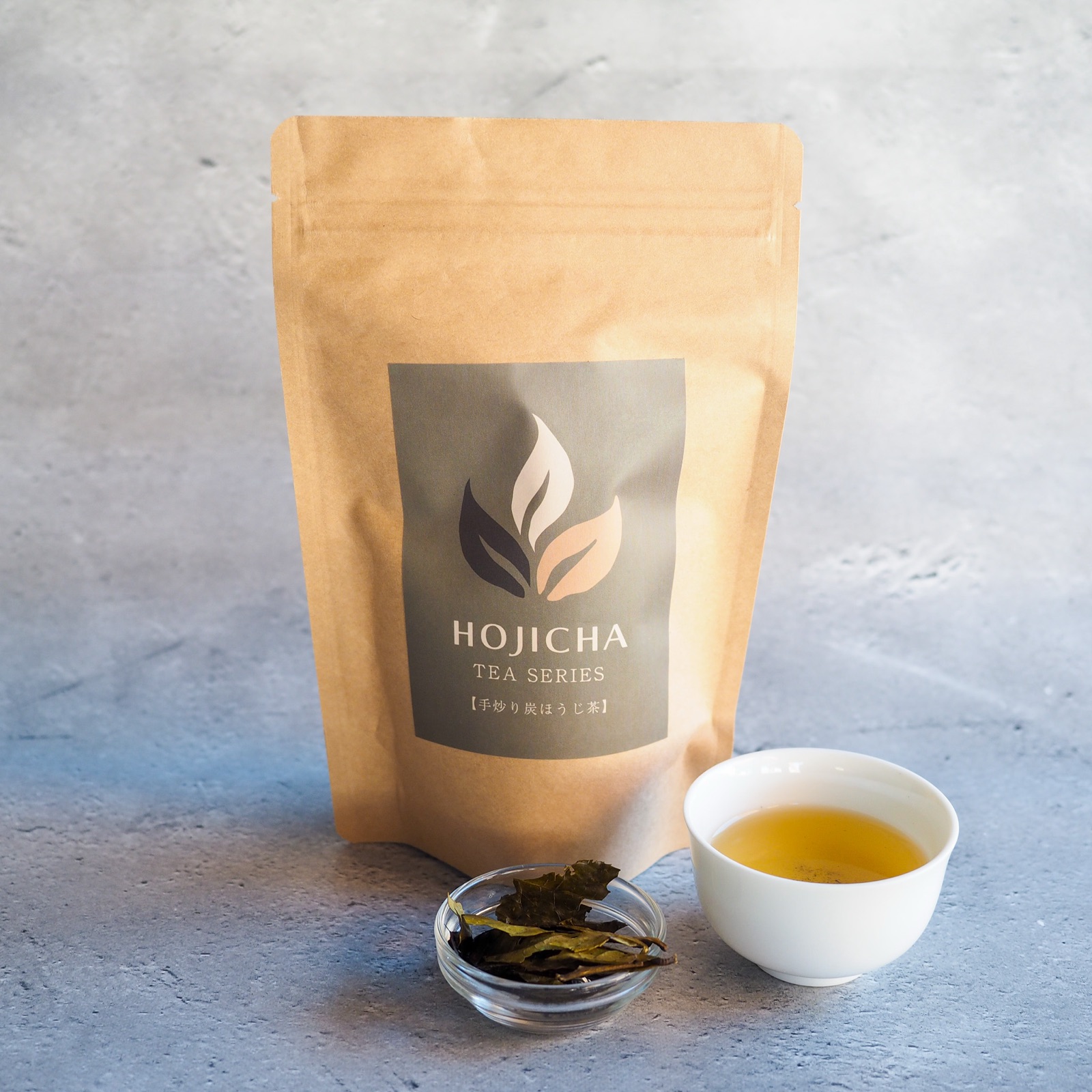 The tea bag package of No.3 Smoky Hand-Roasted Hojicha, with a small glass dish of the dry tea and a white cup of the brewed tea.