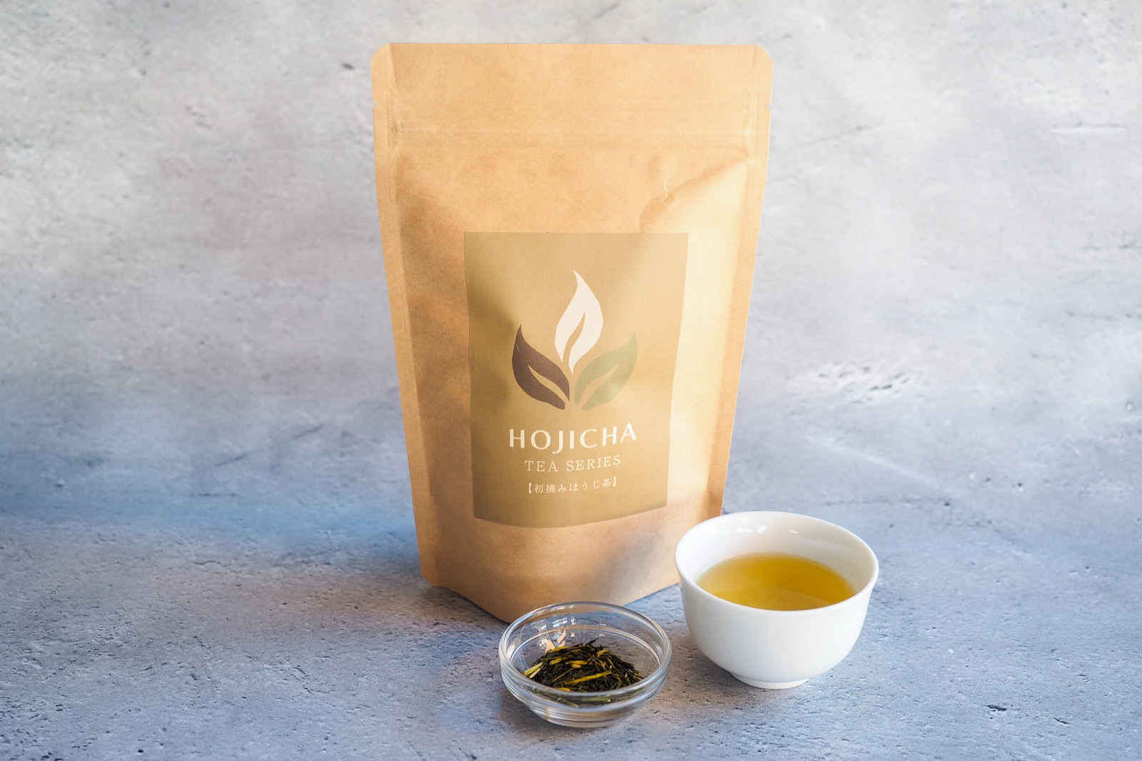 The tea bag package of No.5 First-Pick Hojicha, with a small glass dish of the dry tea and a white cup of the brewed tea.